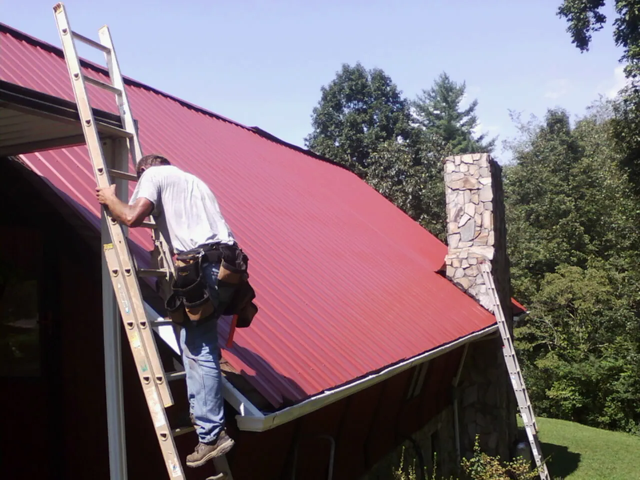 Roofer on a ladder beside a red metal roof and stone chimney — Soffit & Fascia Repair by Roofing Brothers in Levasy